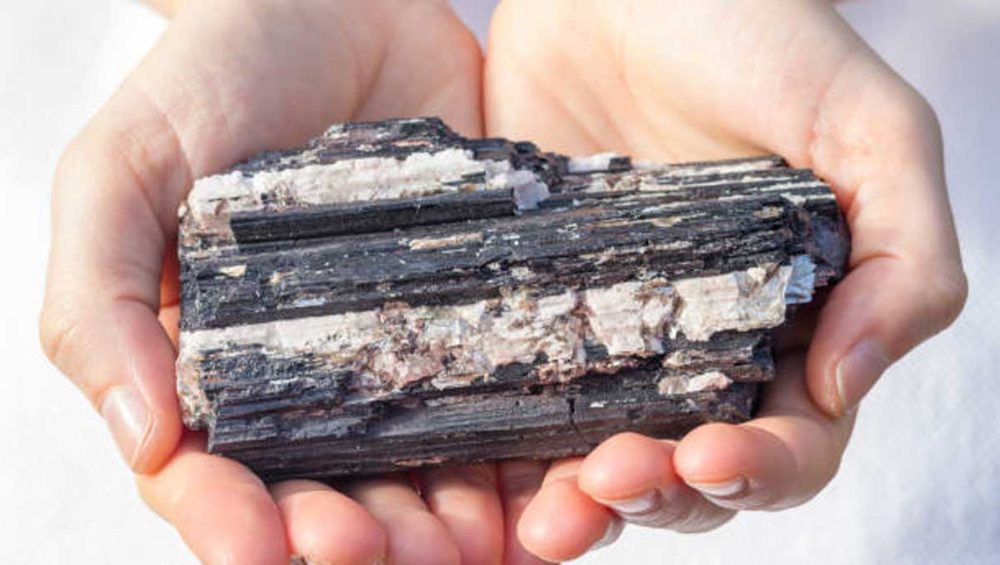 raw black tourmaline crystal held in two hands, showing natural striations and stone matrix