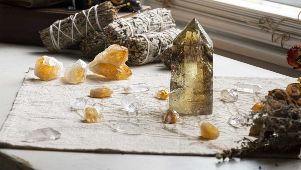 Citrine crystals and clear quartz stones arranged on a cloth beside bundled sage near a window.