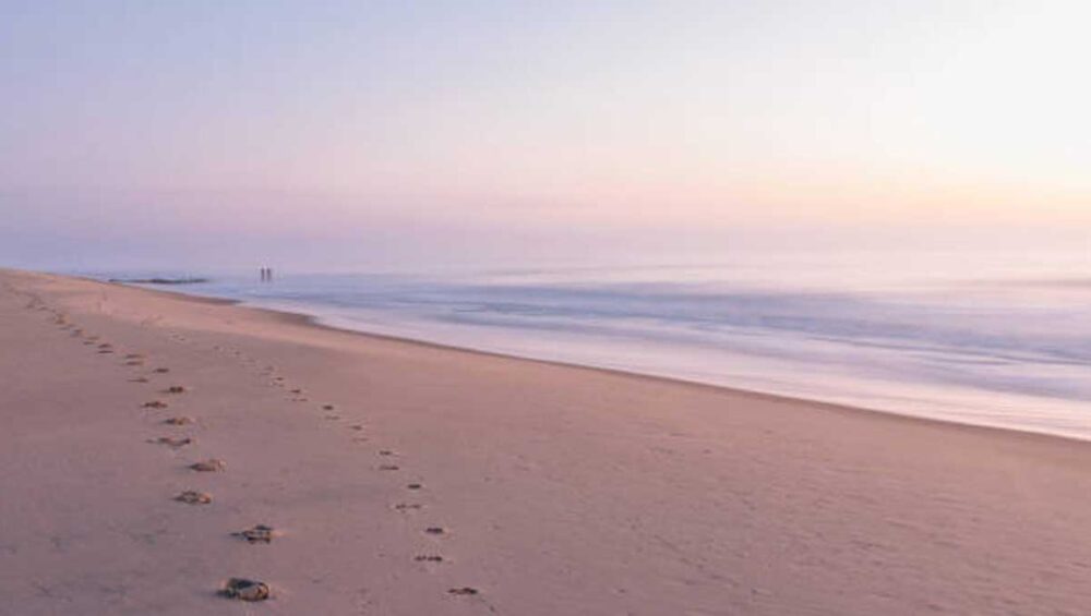 Footprints in sand along a quiet beach at sunrise with soft pastel sky and calm ocean waves