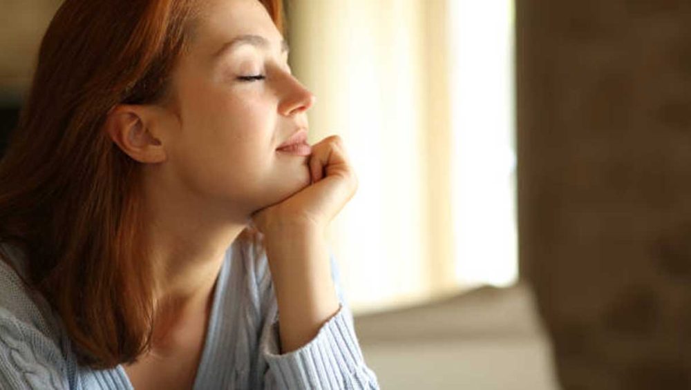 Woman sitting by a window with eyes closed, resting her chin on her hand in soft natural light