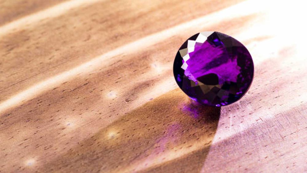 A faceted amethyst birthstone resting on warm wood in natural light.