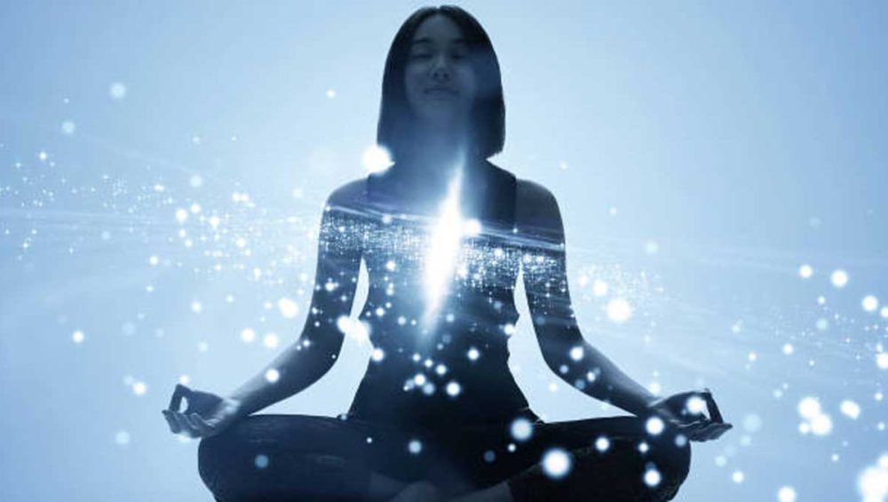Silhouette of a woman meditating with a vertical glowing light along the center of her body against a soft blue background.