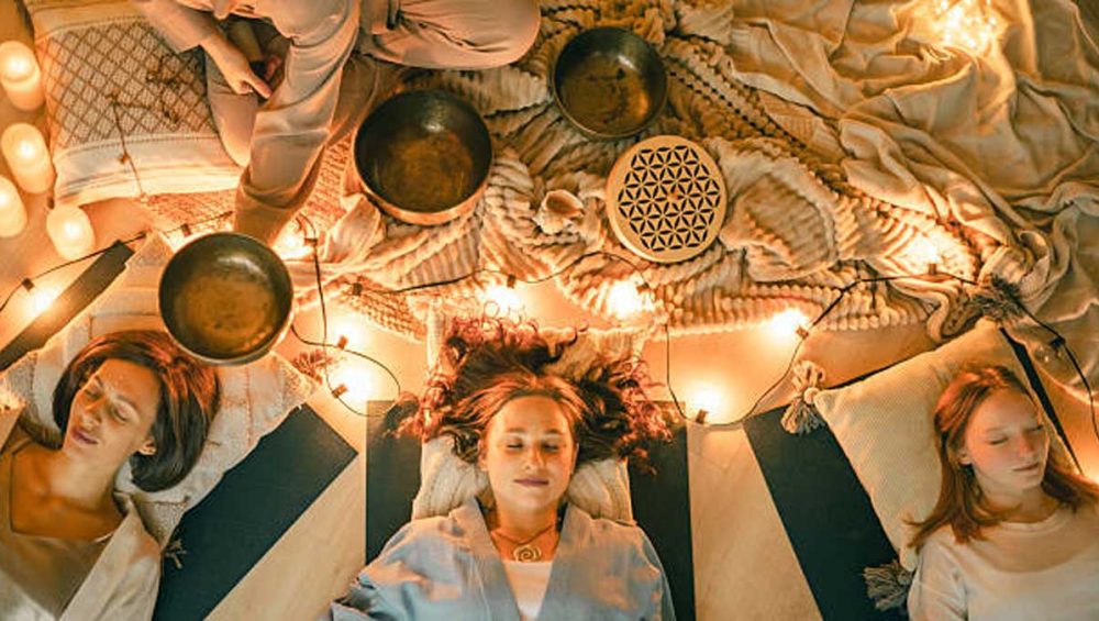 Three women lying on yoga mats during a candlelit sound bath session with singing bowls placed around them.