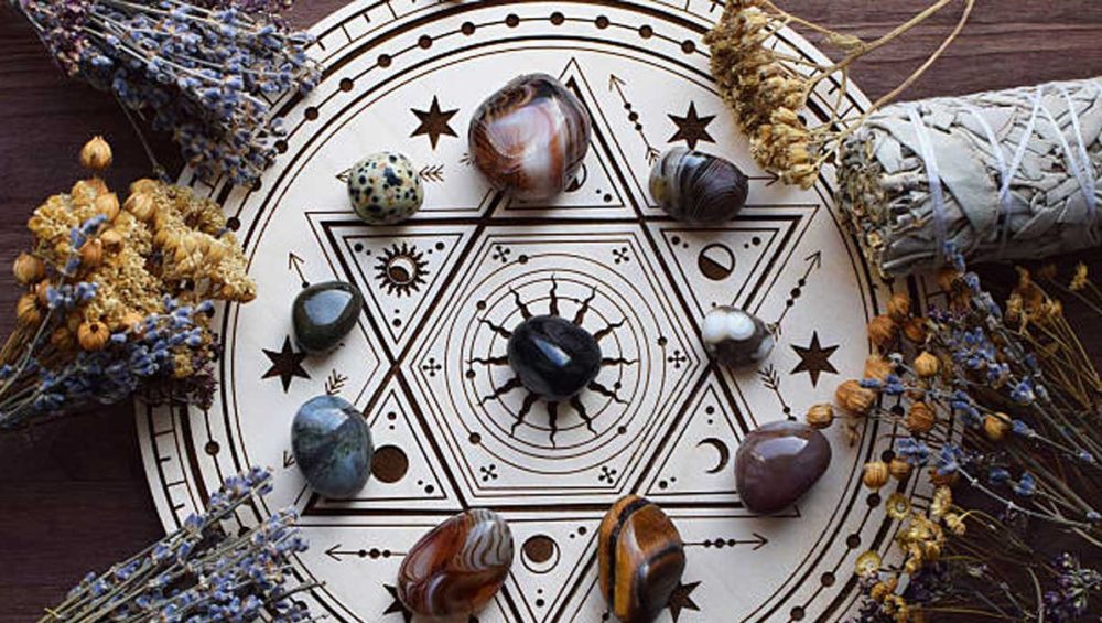 Crystal grid for protection arranged in sacred geometry pattern with black stone at the center, surrounded by polished stones, dried lavender, and sage on a wooden surface.