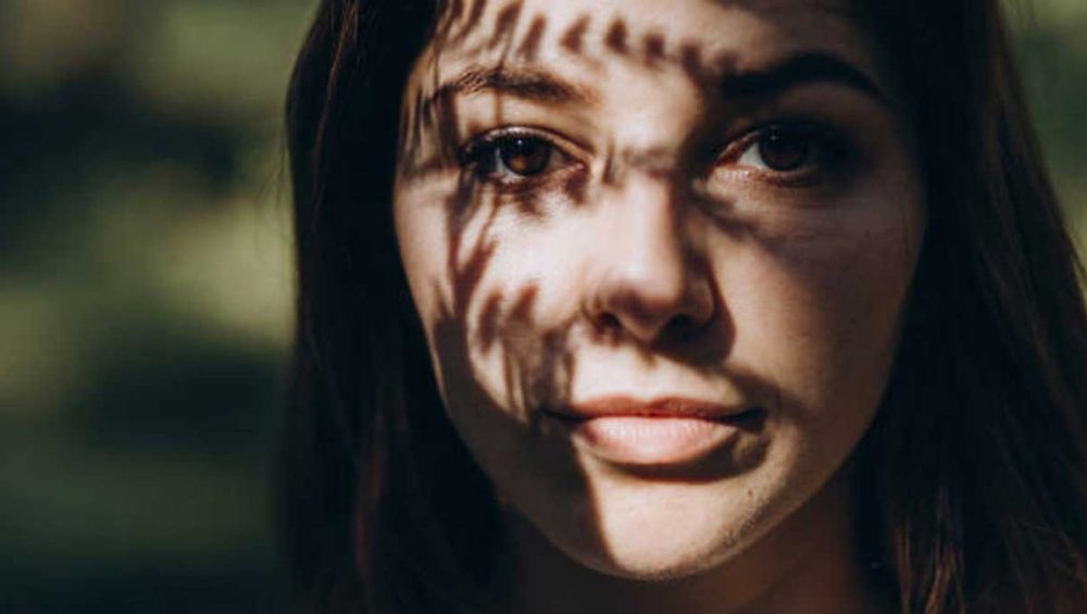 Woman looking directly at the camera with soft shadows from leaves crossing her face.