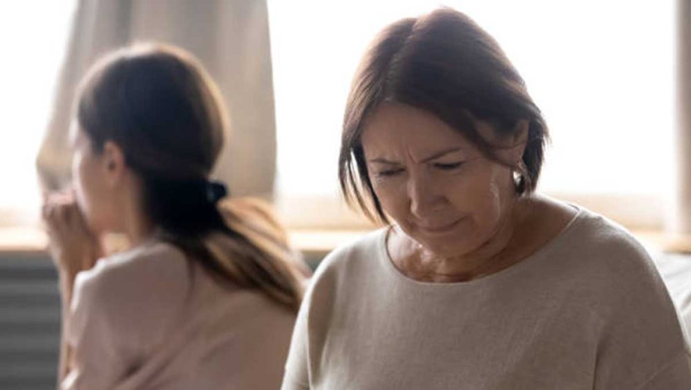 An adult daughter sits turned away while her mother looks down, both appearing emotionally distant.