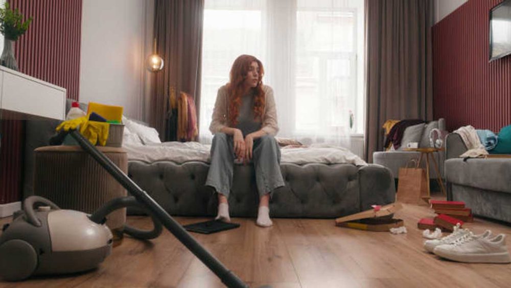 Woman sitting on the edge of a bed in a cluttered room with a vacuum cleaner and scattered household items on the floor