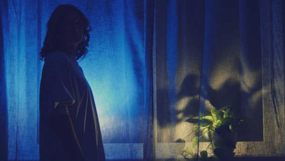 A person standing in silhouette near a softly lit window, surrounded by blue light.