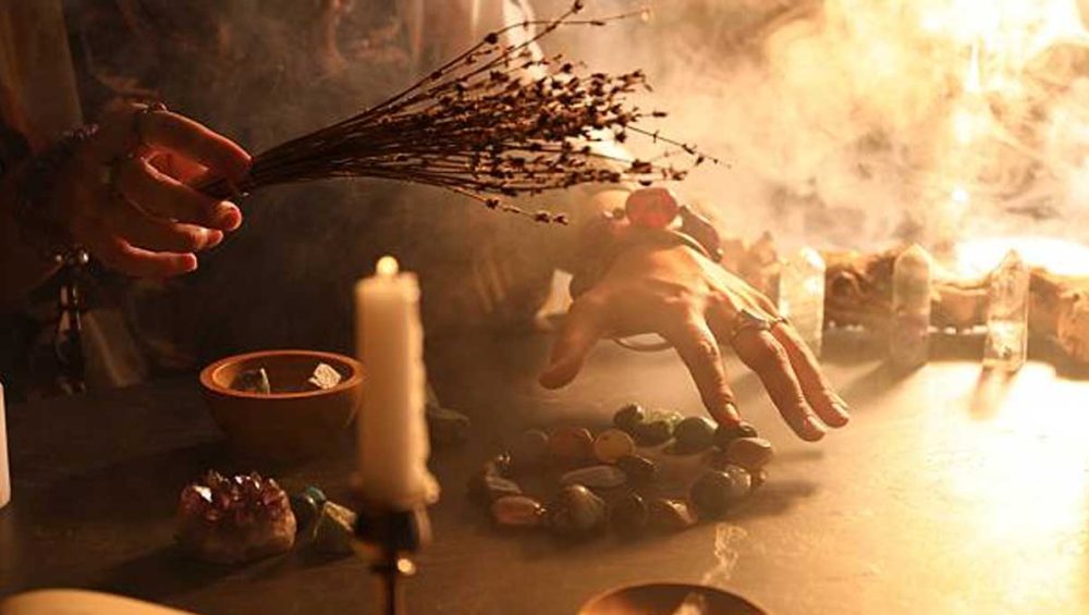 Hand hovering over assorted crystals on a candlelit altar with smoke and dried herbs