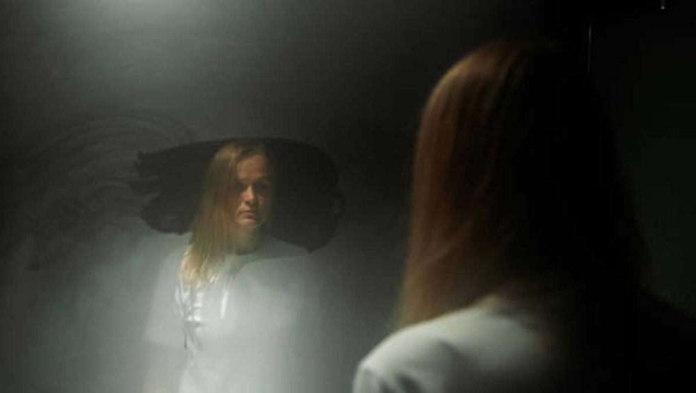 Woman standing in front of a dark mirror with a shadowed, distorted reflection behind her