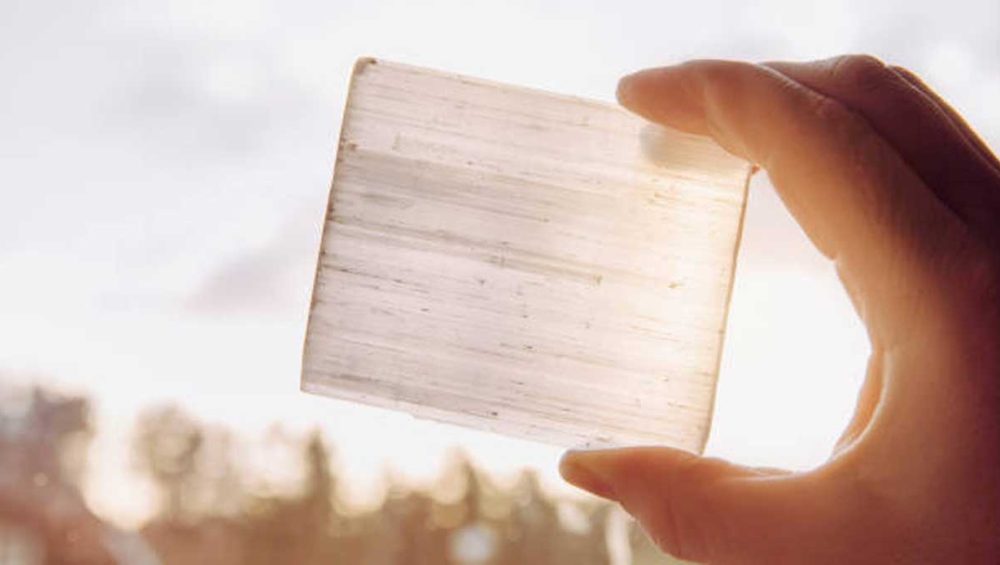 Hand holding a translucent selenite crystal slab up to the light with trees blurred in the background.