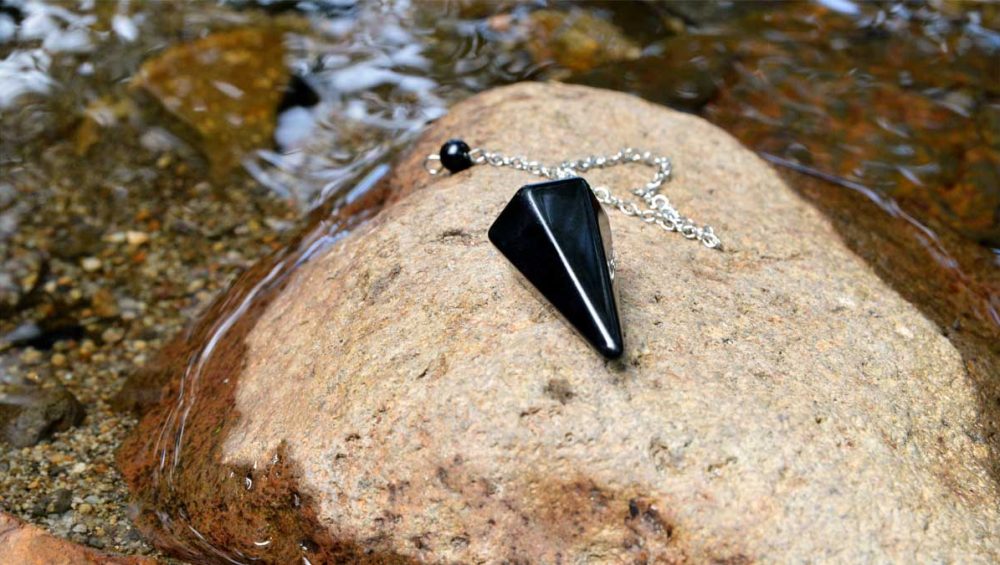 Black onyx pendulum resting on a smooth stone in a shallow flowing stream