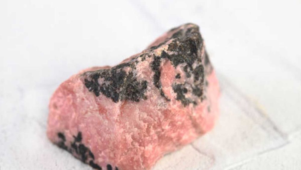 Raw rhodonite stone with pink coloring and black manganese veining on a light background
