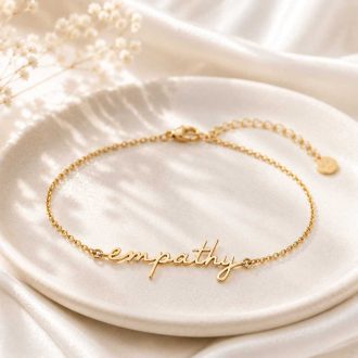 Gold empathy script bracelet displayed on a white ceramic dish with a soft ivory fabric background.