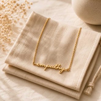 Gold empathy script necklace displayed on a folded linen pouch with a soft ivory background and dried flowers.