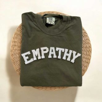 Folded sage green empathy shirt with white varsity-style lettering displayed on a woven rattan tray.
