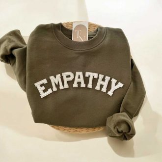 Folded olive green empathy varsity sweatshirt displayed on a woven tray with soft neutral textiles and natural botanical accents.