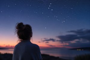 Person looking at a star-filled night sky over calm water during twilight.