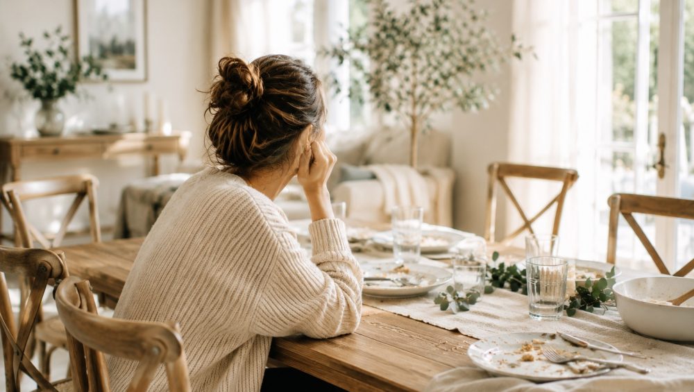 A woman sits alone at a dining table after a meal, quietly looking toward a bright window while empty dishes and glasses remain on the table.