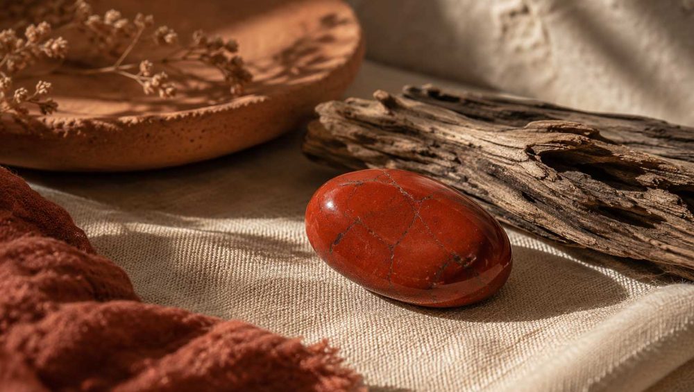 Polished red jasper gemstone resting on neutral linen beside weathered wood and a clay dish in warm natural light.