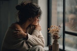 A person sitting quietly by a window hugging themselves while reflecting, with a soft reflection visible in the glass.