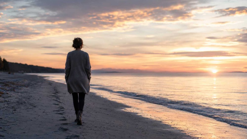 Woman walking alone along a quiet shoreline at sunset