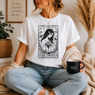 Woman wearing a white T-shirt featuring a black tarot card design titled "The Empath" in a cozy indoor setting.